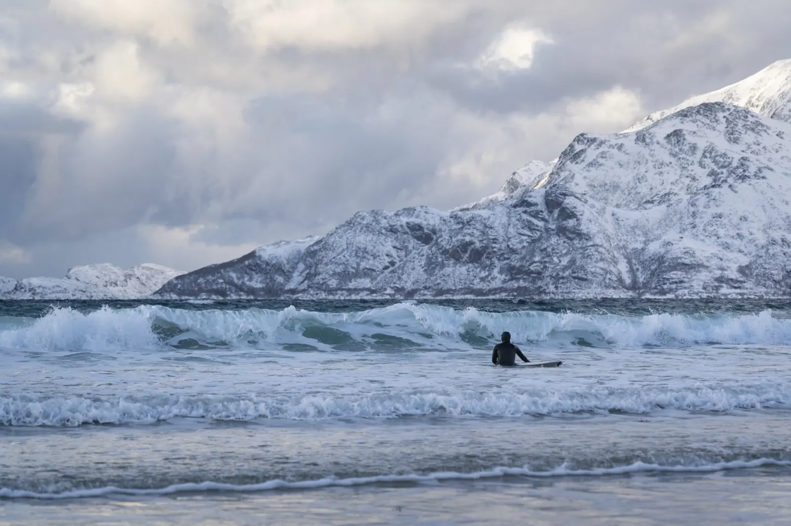 Surfer i bølgene utenfor stranda i Grøtfjord, med snødekte fjell bak
